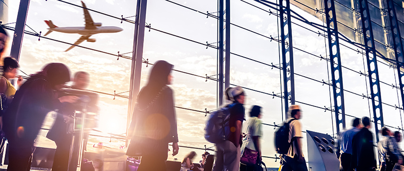 A blurred image of travelers walking through an airport with a plane taking flight in the background.