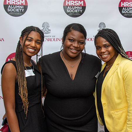 Three people pose for a photo in front of the HRSM Alumni Society backdrop.