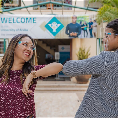 Two students at the University of Aruba wearing clear facial masks bump elbows.