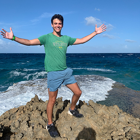 A student stands on a rocky terrain in Aruba with the Caribbean Sea behind.