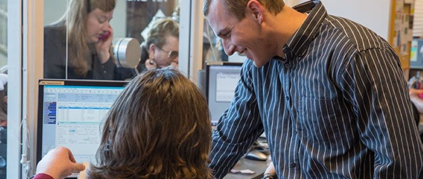 A male employee standing up next to a female employee who is sitting at a desk. They are both looking down some paper. A computer screen is sitting at the desk in the background.