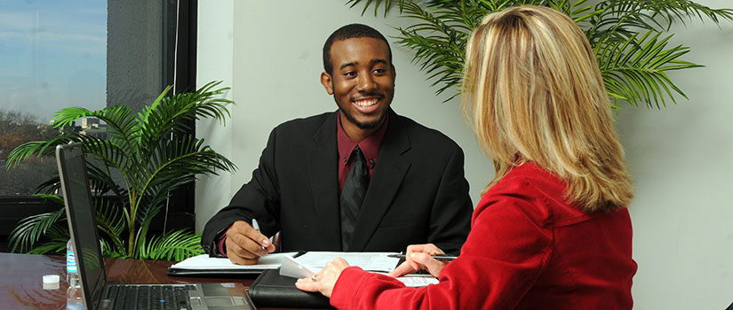 A student meets with a staff member. The two sit at a desk, upon which are papers, a portfolio and a laptop.