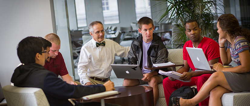 Professor kneeling down to speak to six students who are seated in a circle on armchairs and sofas in a lobby, with their laptops, having a discussion