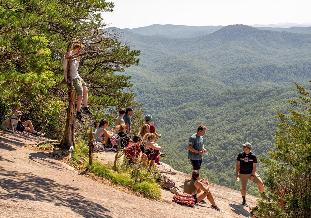 Group of students hanging out on a mountain side overlooking a beautiful view of a valley with mountains in the distance. 