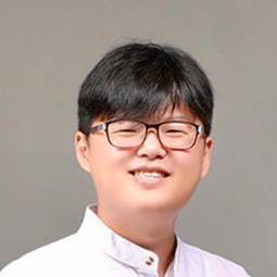 man with dark hair and glasses and white shirt smiling with a tan background 