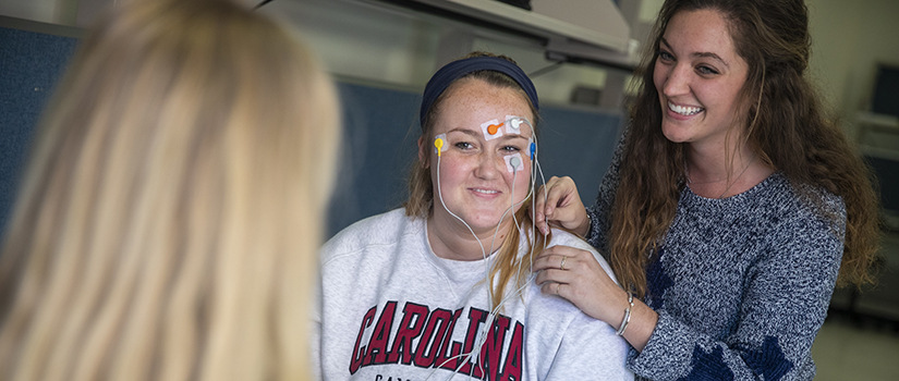 two female students place electrodes on another student's head in a lab