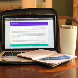 A photo by Joseph Leonard showing his working from home set up. It includes his laptop, a cup, a notebook and a pen.