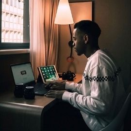 A UofSC student sitting at his desk in a dimly lit room. He has an iPad to his right and his laptop opened in front of him on the Google homepage