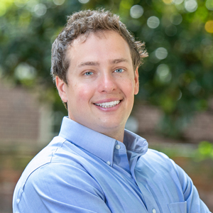 Sam McQuillin, wearing a blue button-up, stands on campus